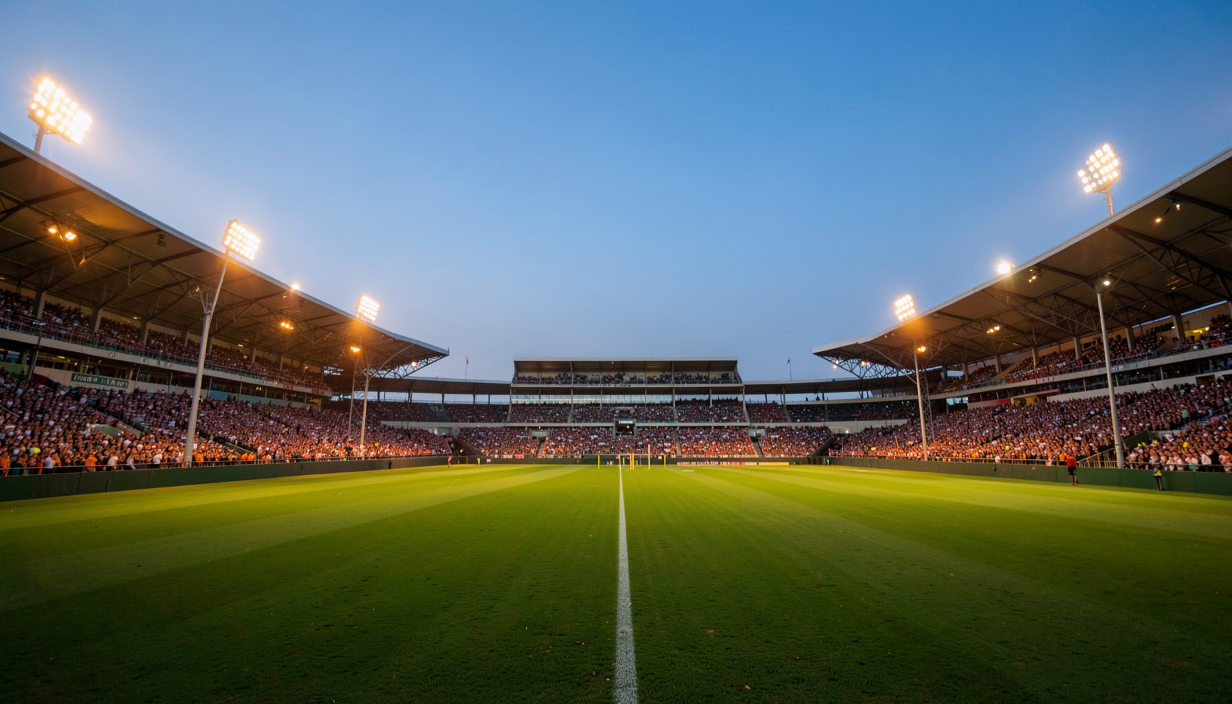 A wide atmospheric shot of a cricket stadium at dusk, warm amber floodlights illuminating the green field, with a soft blue sky fading into evening