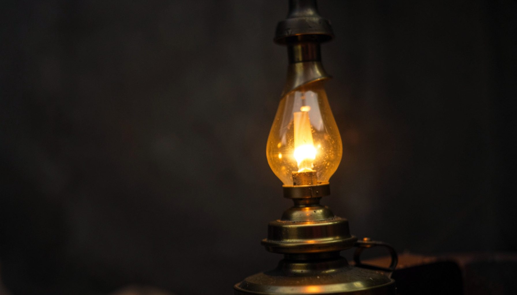 A close-up of a traditional brass lamp glowing warmly against a dark, muted background, conveying a sense of heritage and thoughtful correspondence.